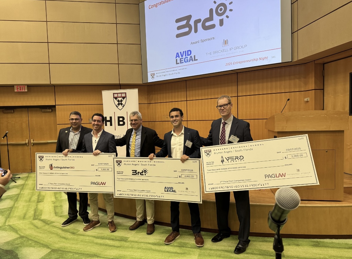 Dillon Abend holding the first place 3rd-i award check at the Harvard Alumni Angels South Florida pitch competition, with the 3rd-i logo on the screen behind him